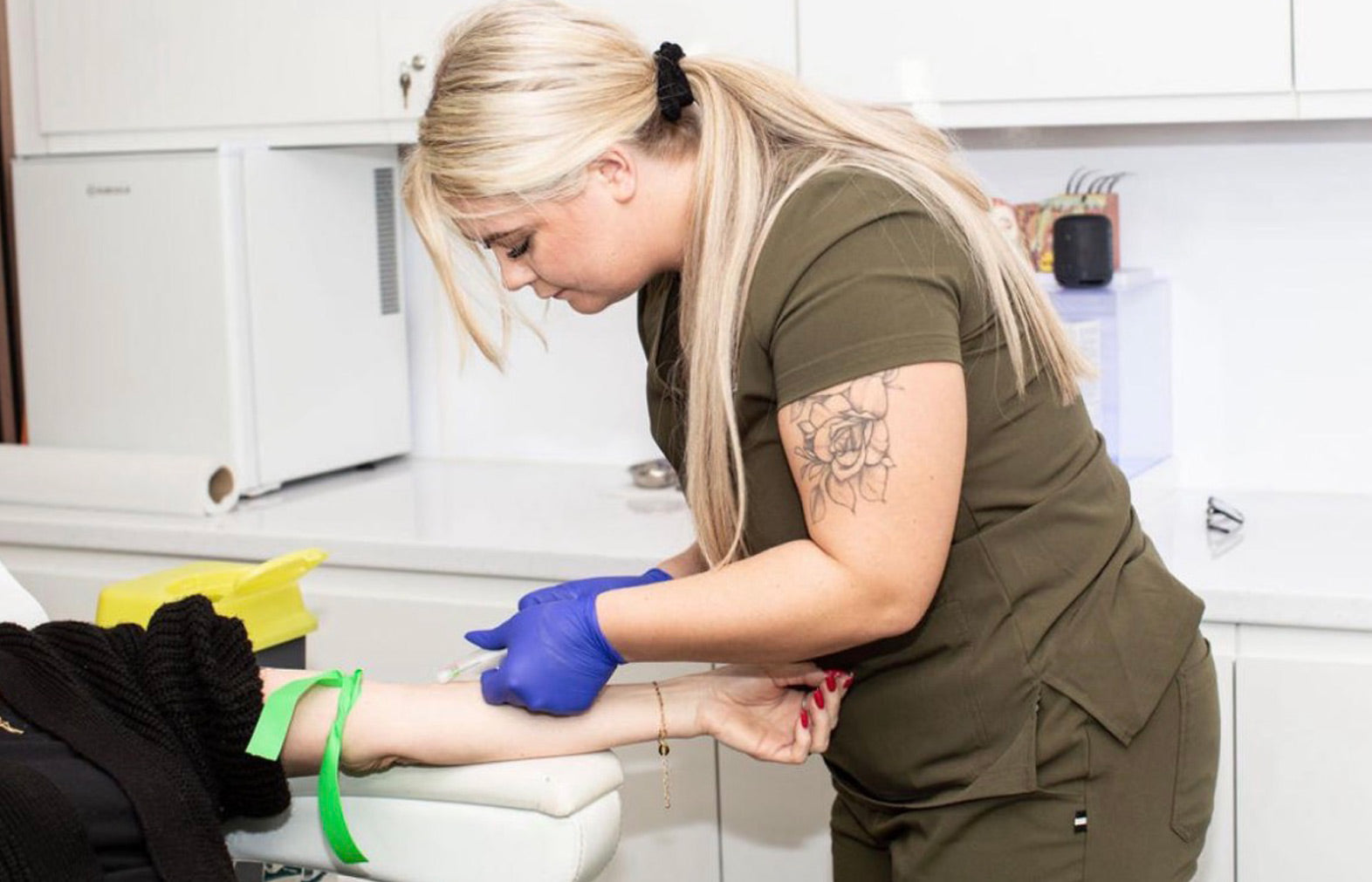 Person in green uniform with gloves assisting another person in a kitchen setting